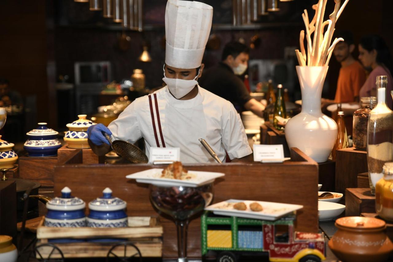 Hotel Radisson Blu Plaza Delhi Airport Nuova Delhi Esterno foto The photo depicts a chef in a kitchen setting. He is wearing a white chef's uniform and a tall white hat, along with gloves. The chef appears to be preparing food, possibly serving from a buffet or a display area. Various dishes are arranged on a woo