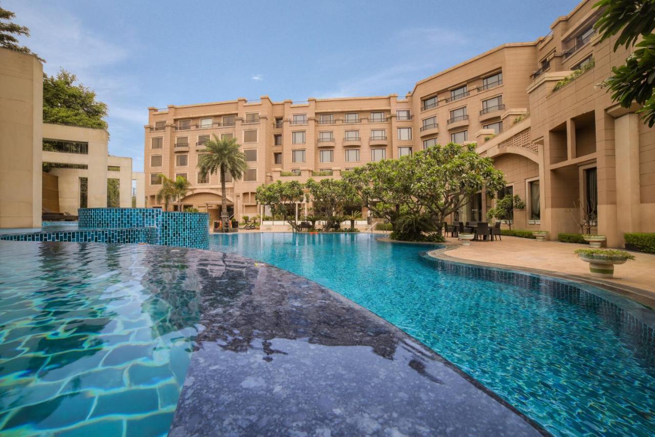 Hotel Radisson Blu Plaza Delhi Airport Nuova Delhi Esterno foto The photo shows a luxurious pool area with a large swimming pool in the foreground. The pool has a smooth, blue surface and features a dark stone edge. Surrounding the pool, there are well-maintained gardens with palm trees and tropical plants. In th