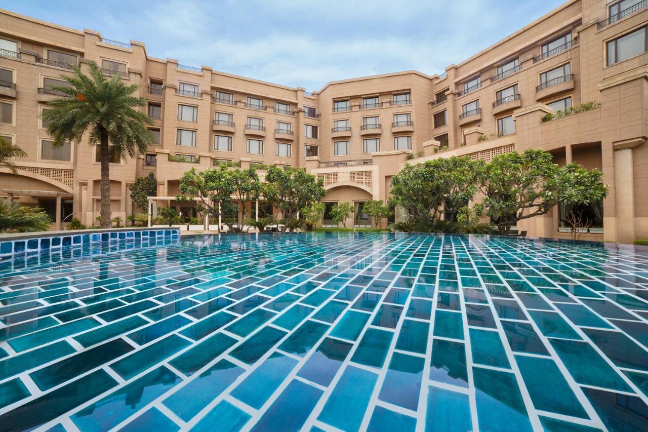 Hotel Radisson Blu Plaza Delhi Airport Nuova Delhi Esterno foto The photo shows a large swimming pool with a distinctive blue tiled surface, reflecting shades of light and water. Surrounding the pool is a landscaped area featuring greenery, including trees and shrubs. In the background, there is a stylish buildin