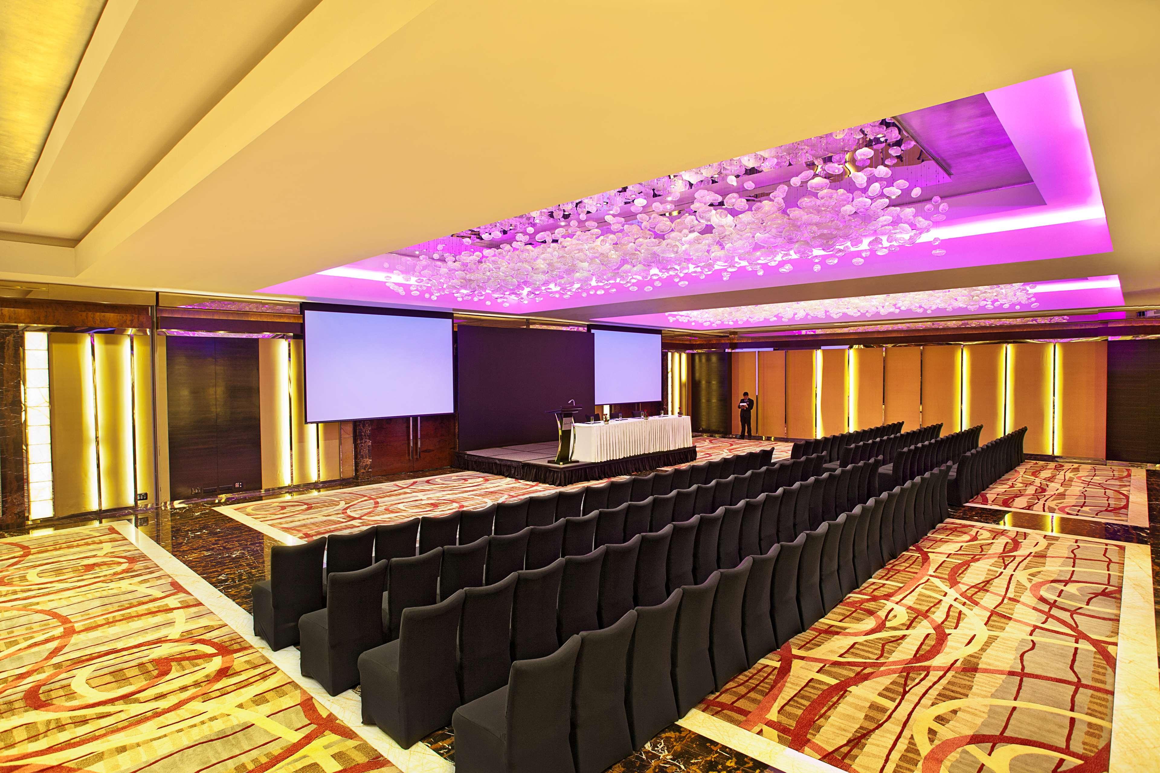 Hotel Radisson Blu Plaza Delhi Airport Nuova Delhi Esterno foto The photo shows a spacious conference room set up for an event. There are rows of black chairs arranged in front of a stage area. The stage features a table with items placed on it, and two large screens are positioned on either side. The room has mo