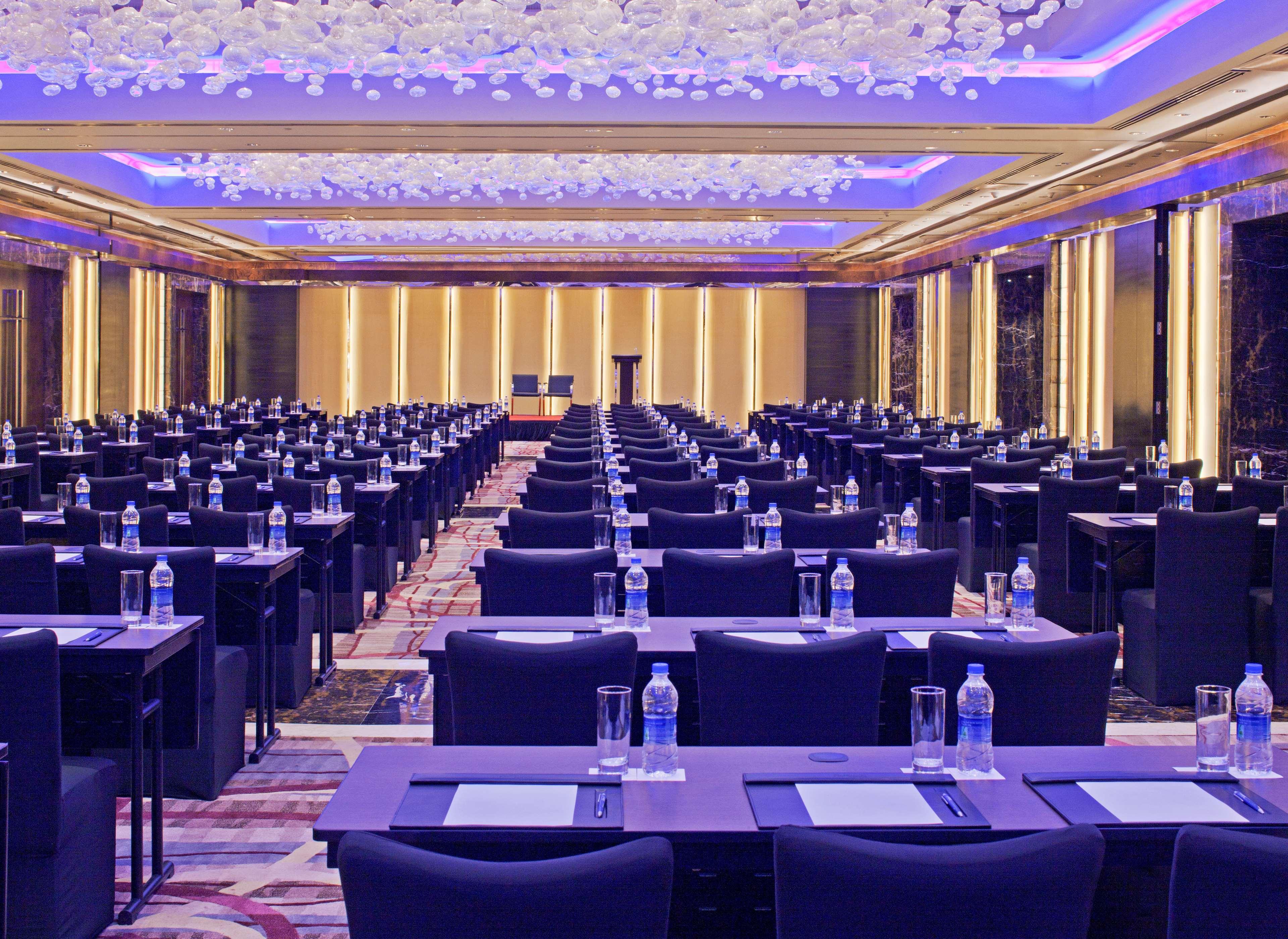 Hotel Radisson Blu Plaza Delhi Airport Nuova Delhi Esterno foto The photo shows a large conference room arranged for a meeting or seminar. There are many rows of rectangular tables, each paired with chairs, facing a stage or presentation area at the front. The tables have water bottles and notepads on them, indic