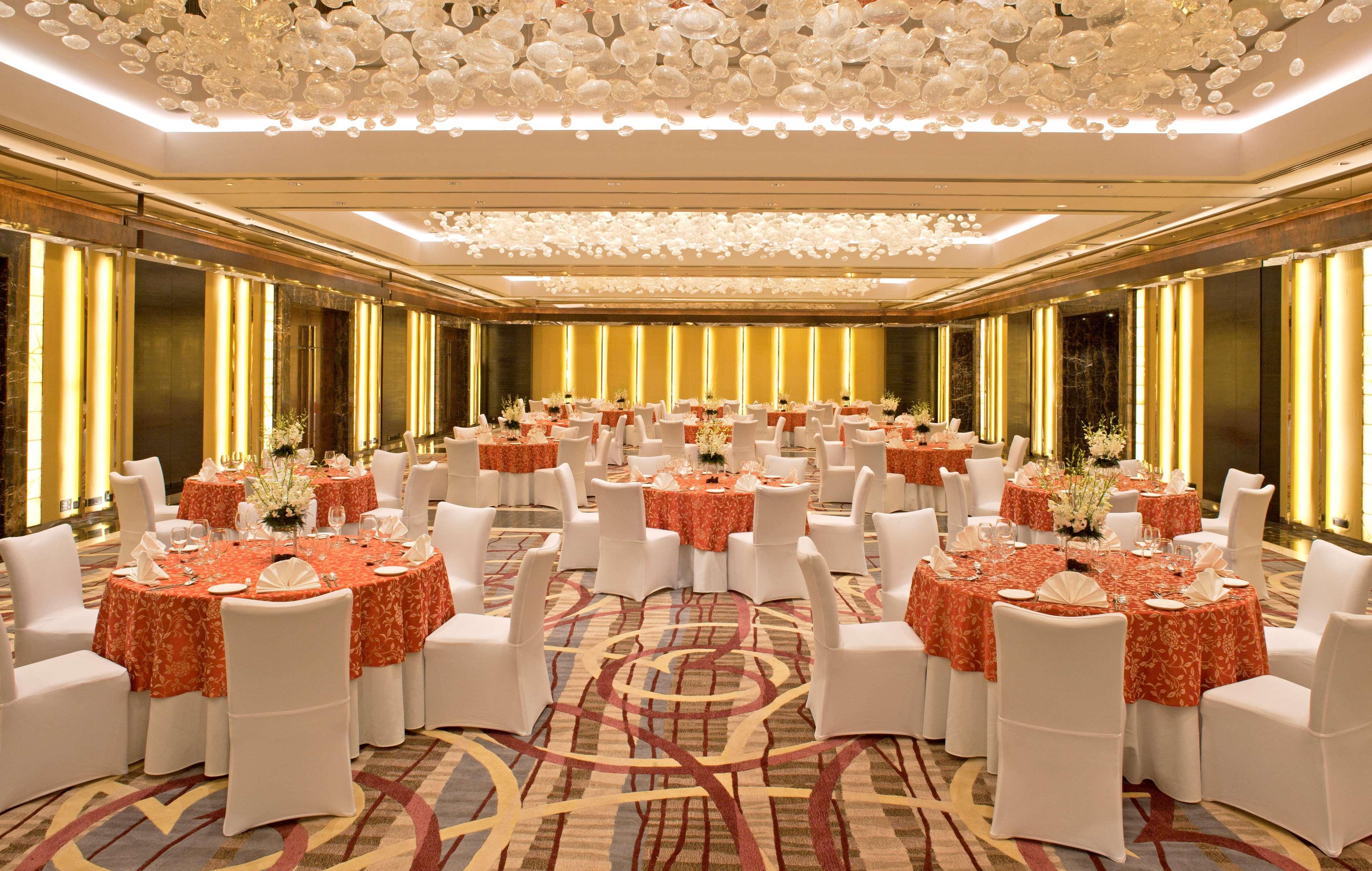 Hotel Radisson Blu Plaza Delhi Airport Nuova Delhi Esterno foto The photo showcases an elegantly arranged banquet hall. The room features multiple round tables covered with orange tablecloths, each adorned with floral centerpieces. White chairs surround the tables. The space is well-lit with soft lighting, and th