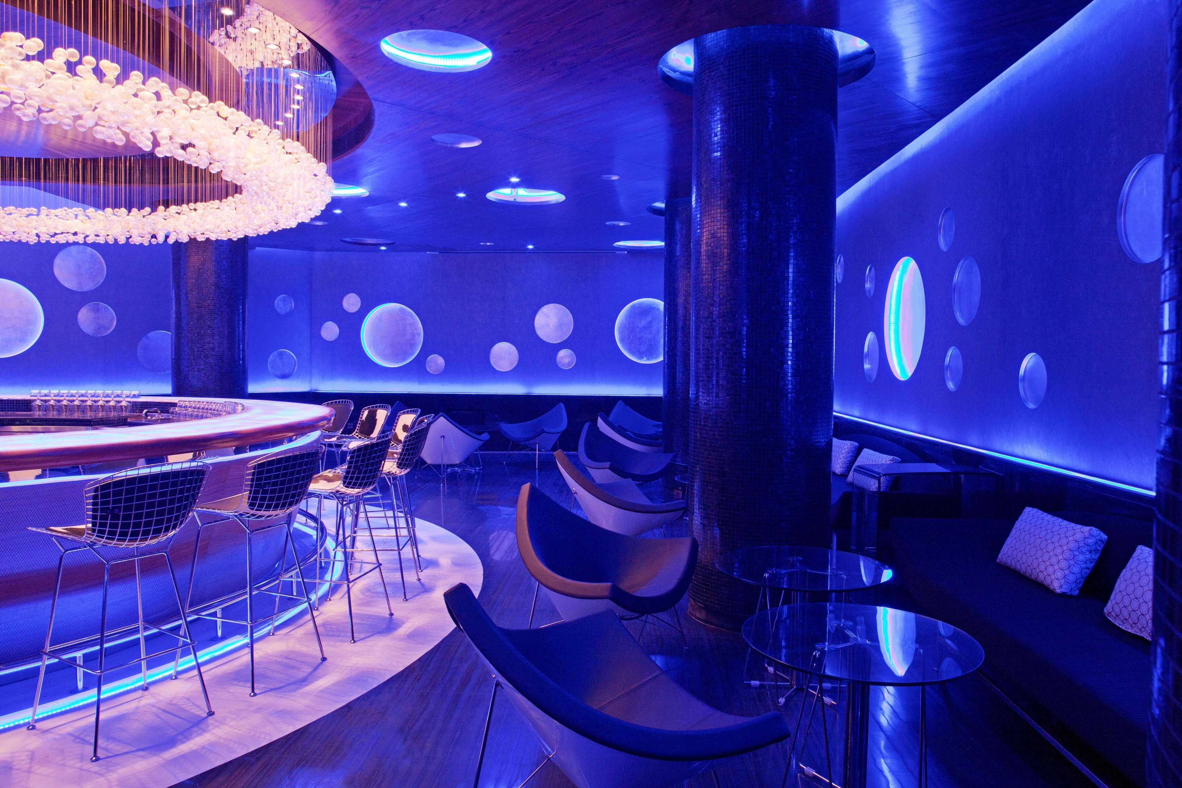 Hotel Radisson Blu Plaza Delhi Airport Nuova Delhi Esterno foto The photo showcases a modern and stylish bar or lounge area. The interior features a predominantly blue color scheme, with soft lighting that creates a cozy and atmospheric ambiance. 

A large circular bar is prominent in the center, illuminated with