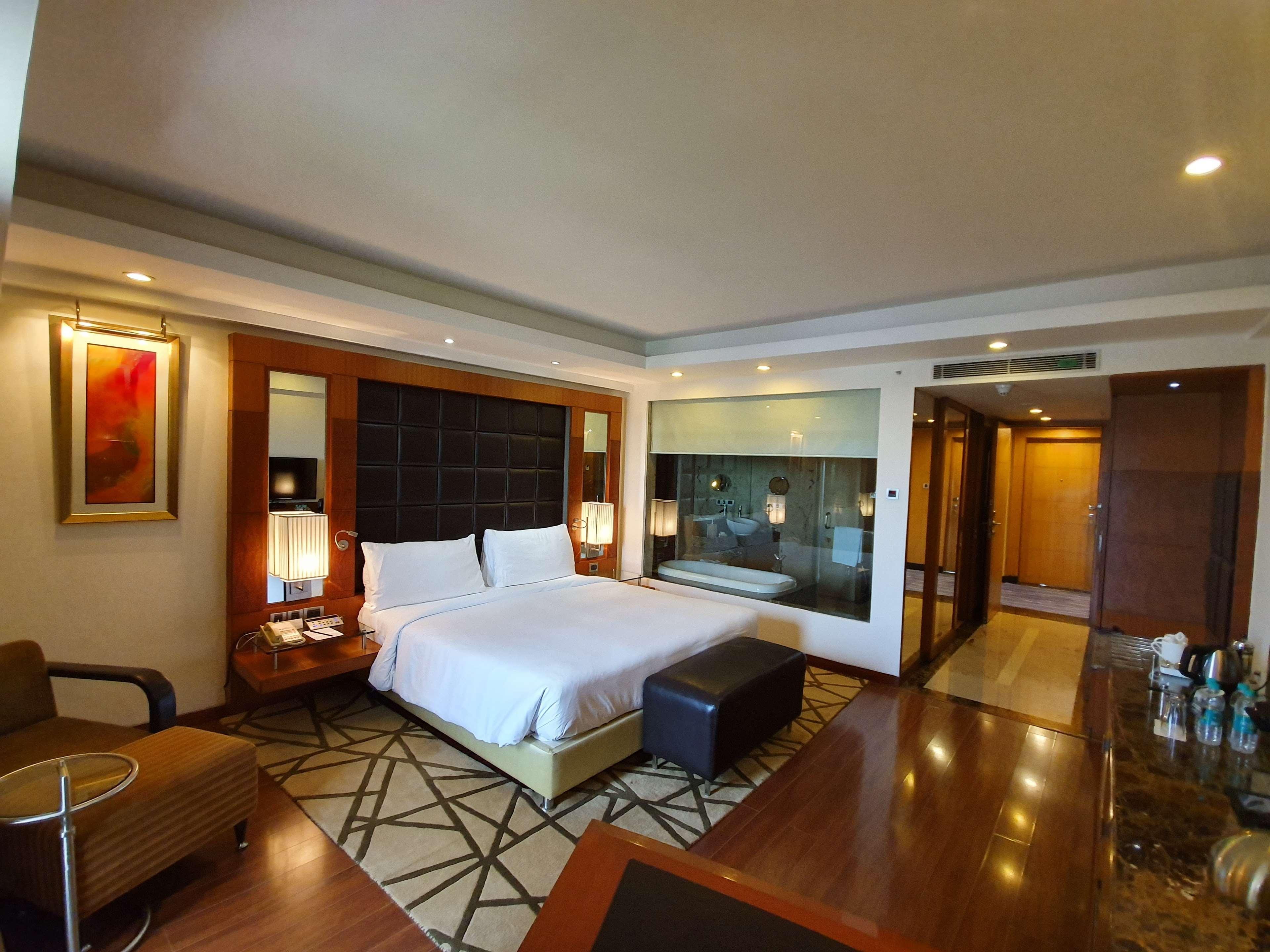 Hotel Radisson Blu Plaza Delhi Airport Nuova Delhi Esterno foto The photo shows a modern hotel room featuring a large bed with white bedding, positioned against a stylish headboard. There are two bedside lamps on either side of the bed. A comfortable armchair is placed in one corner, along with a small side table