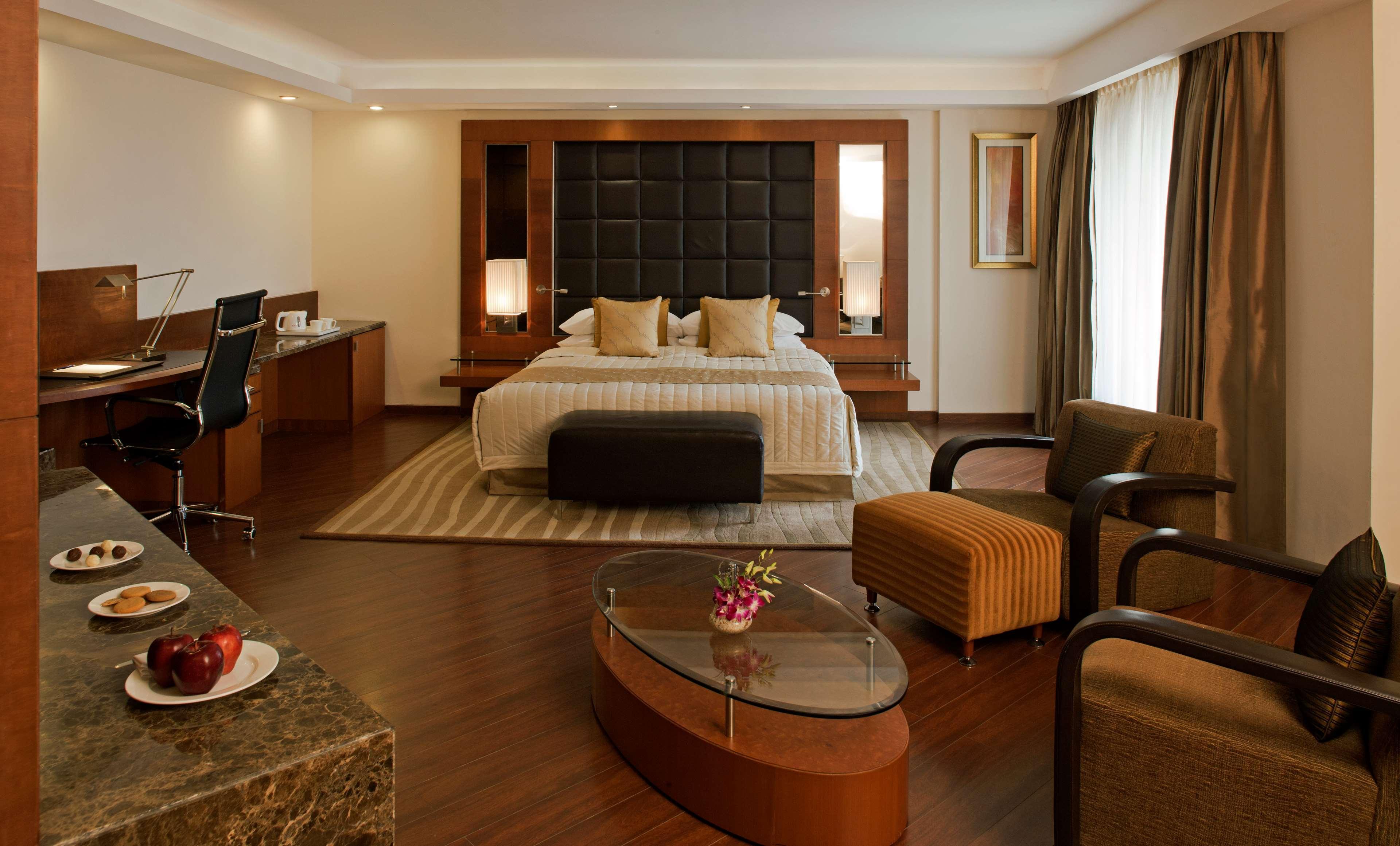 Hotel Radisson Blu Plaza Delhi Airport Nuova Delhi Esterno foto The photo shows a modern hotel room with a stylish and sophisticated design. In the center is a bed with a dark headboard, complemented by two decorative pillows. Nearby, there is a small, dark bench at the foot of the bed. 

To one side, there is a 