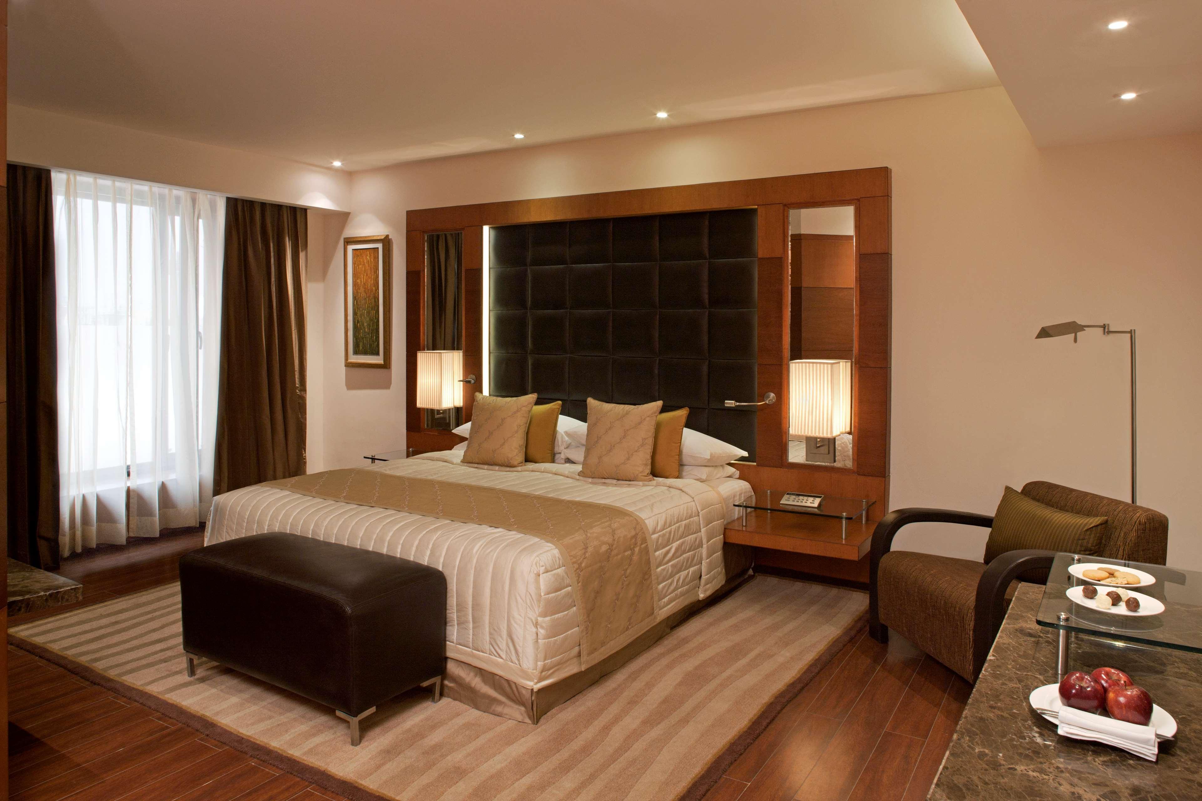 Hotel Radisson Blu Plaza Delhi Airport Nuova Delhi Esterno foto The photo showcases a modern hotel room. It features a large bed with a cream and beige color scheme, adorned with several cushions. Behind the bed is a stylish headboard with a black tufted design. On either side of the bed, there are contemporary l