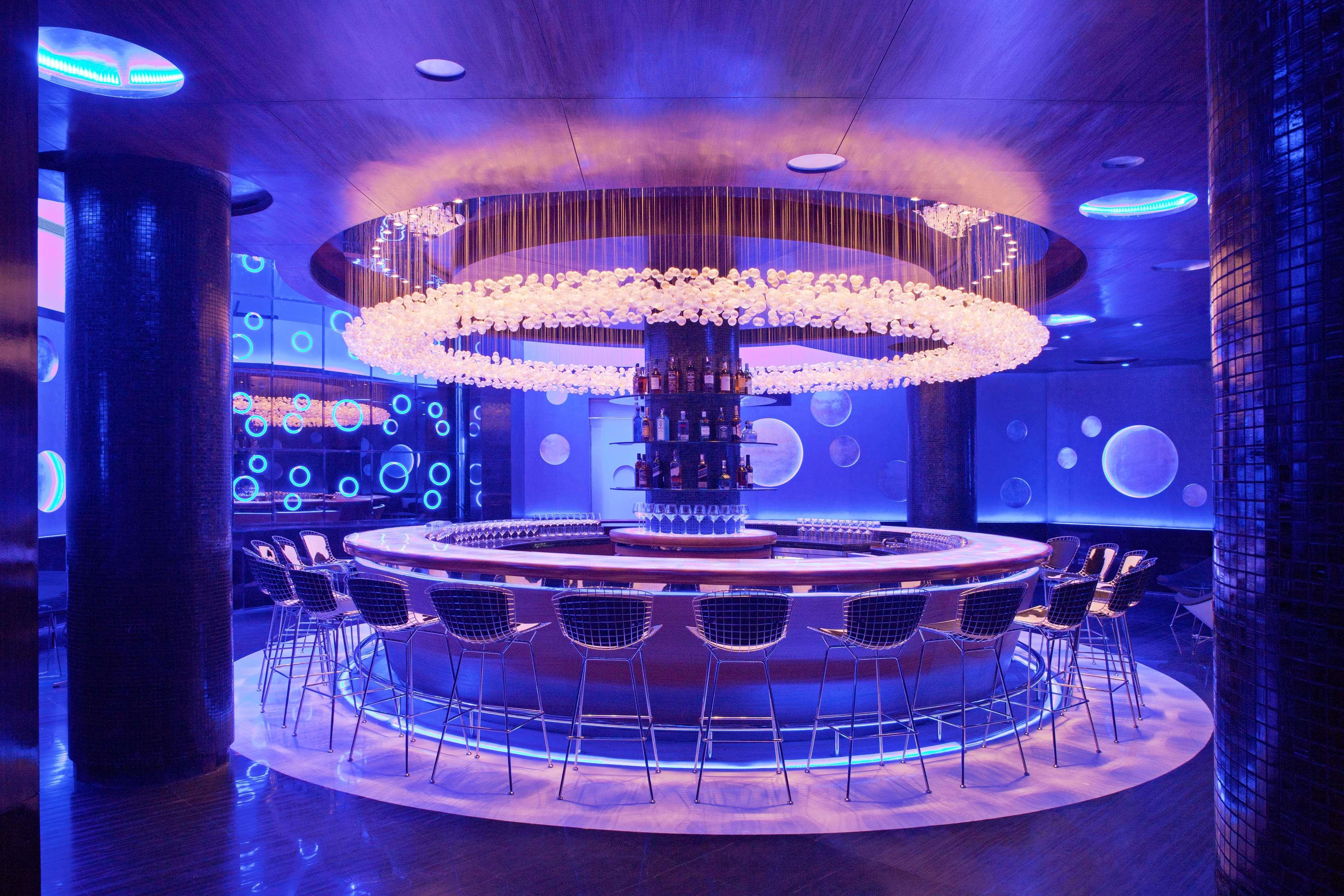 Hotel Radisson Blu Plaza Delhi Airport Nuova Delhi Esterno foto The photo shows a modern and stylish bar area with a circular bar counter at the center. The bar is illuminated by a striking chandelier made of translucent materials, creating a glowing effect. The surrounding space has a sleek design with blue ligh