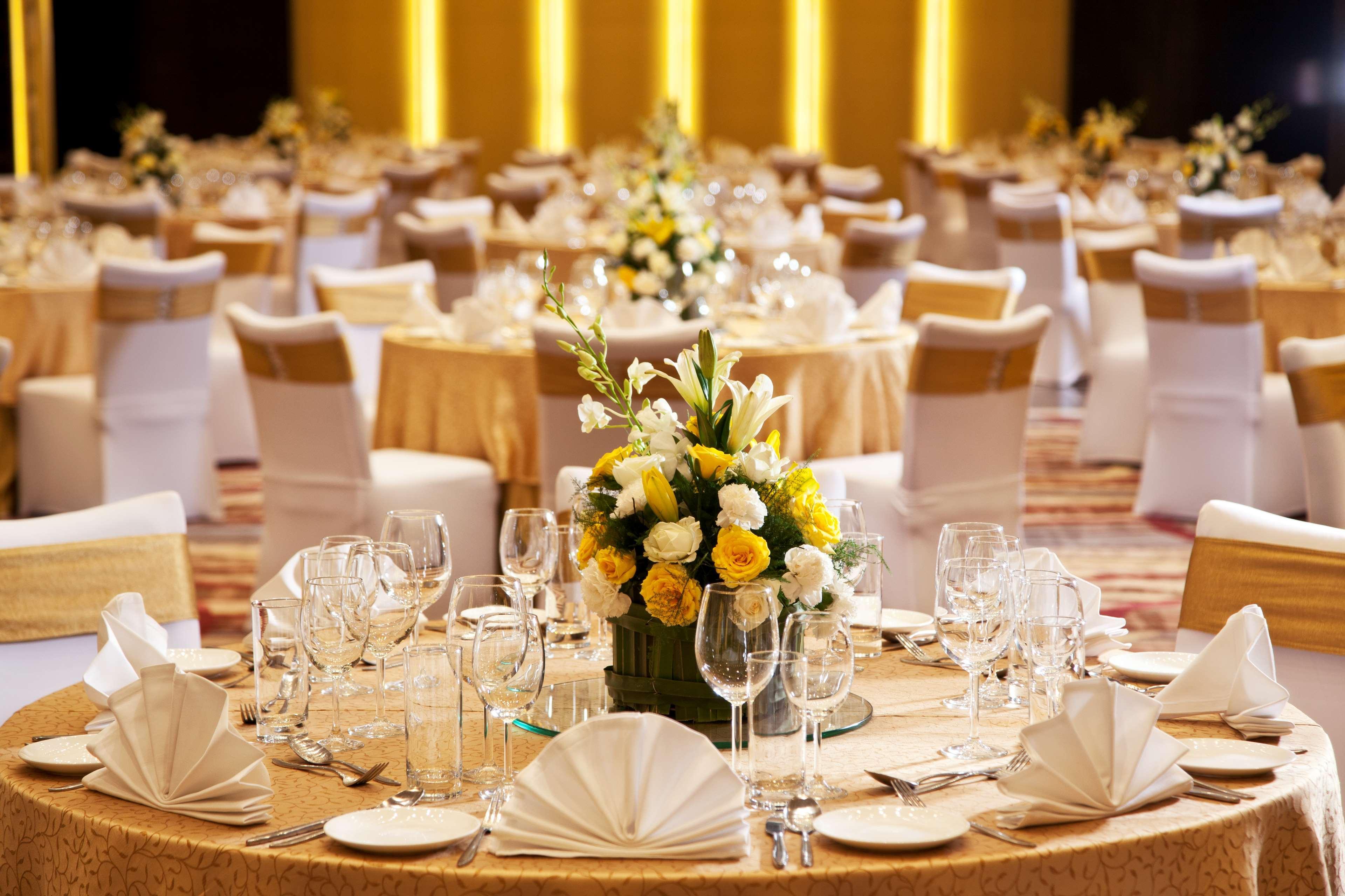 Hotel Radisson Blu Plaza Delhi Airport Nuova Delhi Esterno foto The photo shows a beautifully arranged dining setup in a banquet or event hall. Several tables are decorated with elegant tablecloths, and each table features a centerpiece made up of flowers, including yellow and white blooms. There are neatly arran