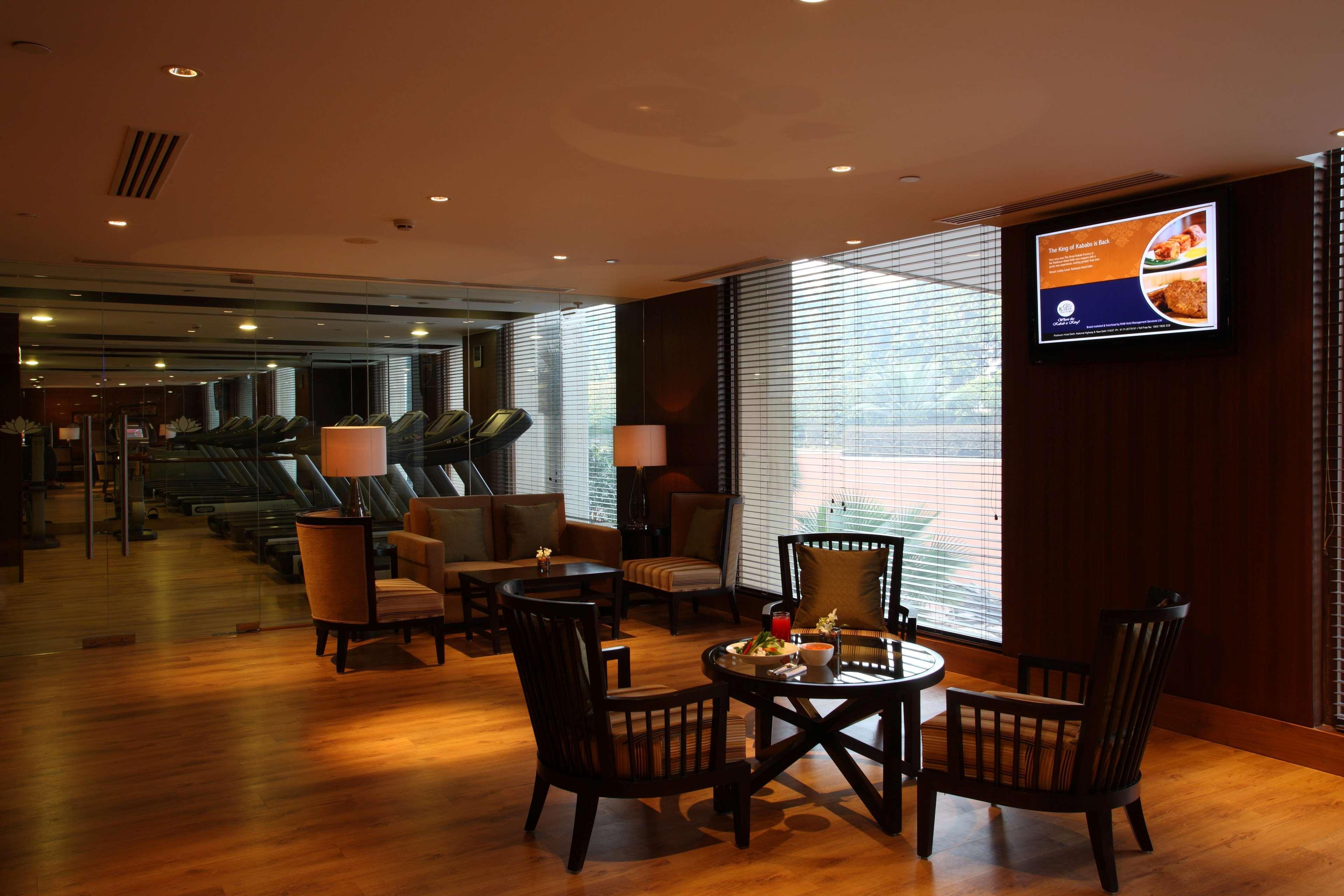 Hotel Radisson Blu Plaza Delhi Airport Nuova Delhi Esterno foto The photo shows a modern interior space, likely a lounge or waiting area. It features several comfortable seating arrangements with wooden chairs and a table in the center. The lighting is warm and inviting, created by ceiling lights and table lamps.