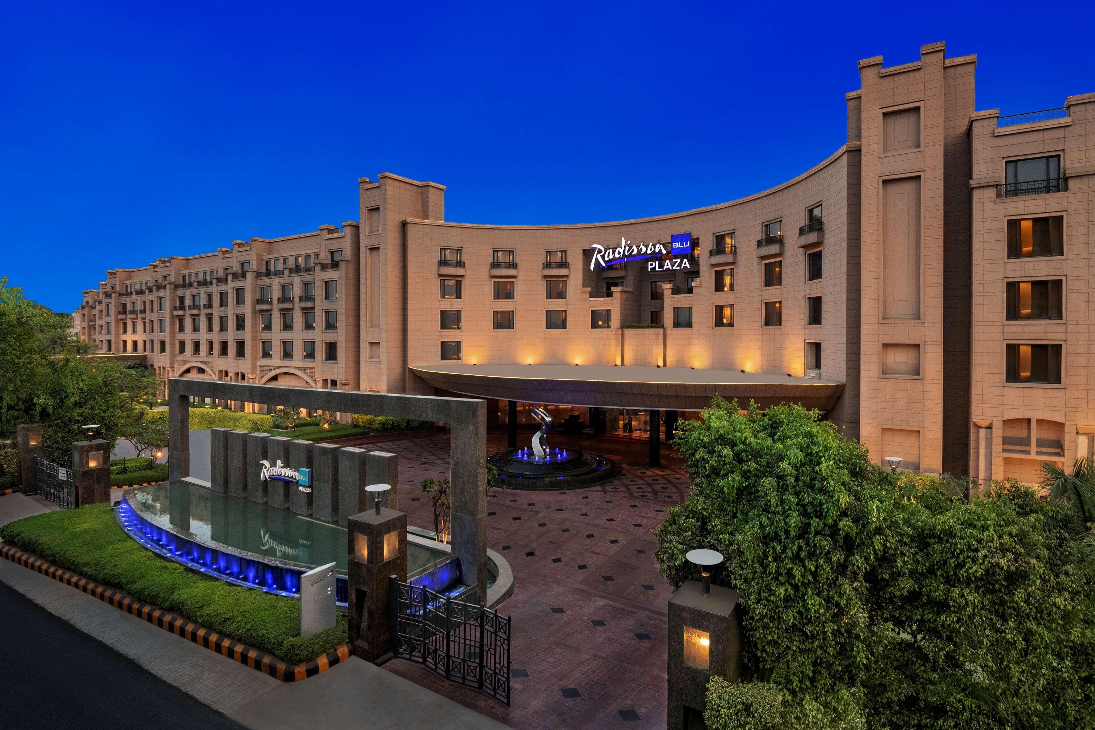 Hotel Radisson Blu Plaza Delhi Airport Nuova Delhi Esterno foto The photo shows the exterior of a Radisson Plaza hotel, featuring a modern architectural design. The building is well-lit against a twilight sky, creating an inviting atmosphere. In the foreground, there is a water feature, likely a small fountain, s