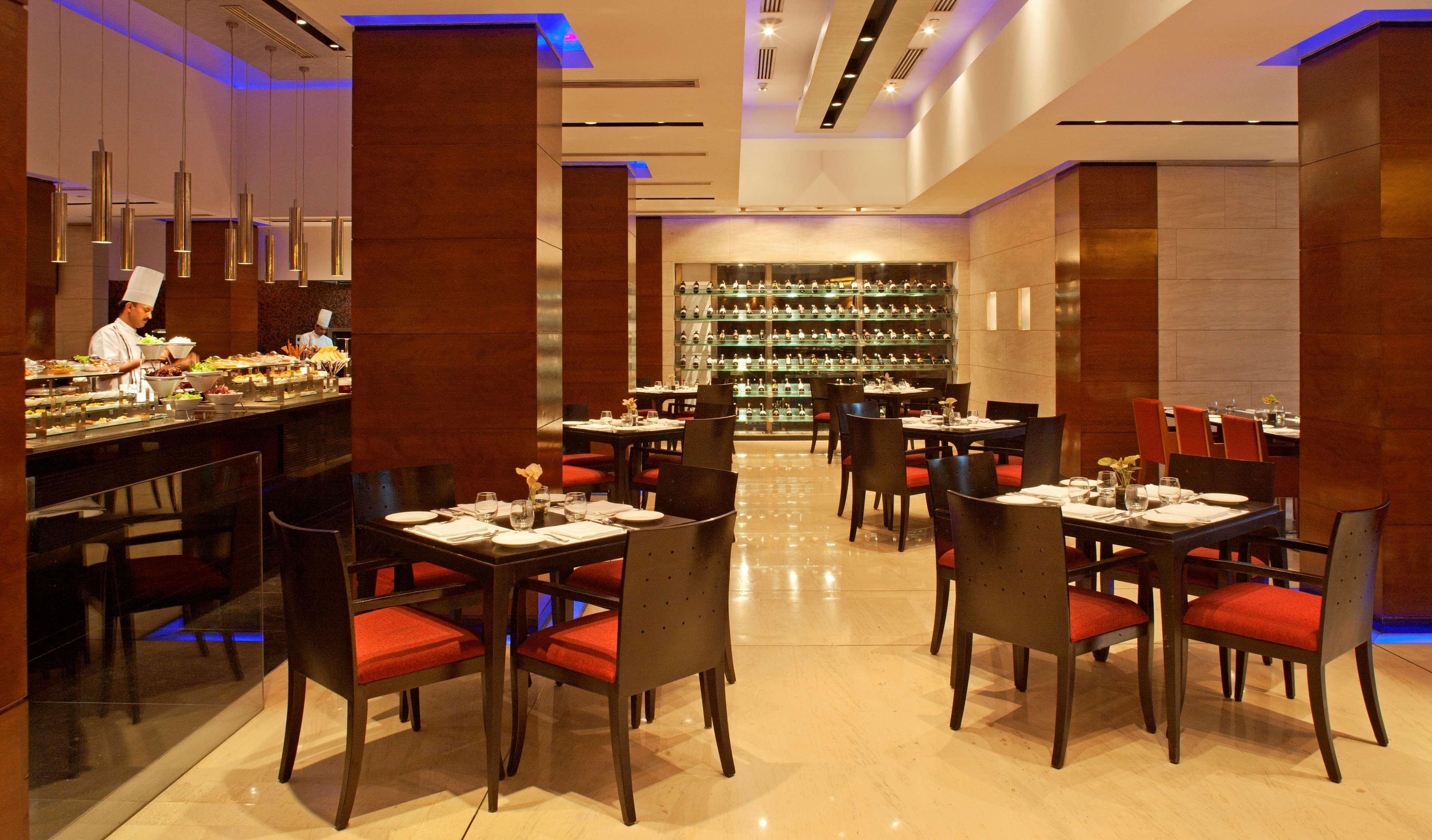 Hotel Radisson Blu Plaza Delhi Airport Nuova Delhi Esterno foto The image depicts a modern restaurant interior. It features a spacious dining area with stylish wooden furniture, including tables with black frames and red seats. The atmosphere is enhanced by soft lighting, with blue accents on the ceiling. A buffe