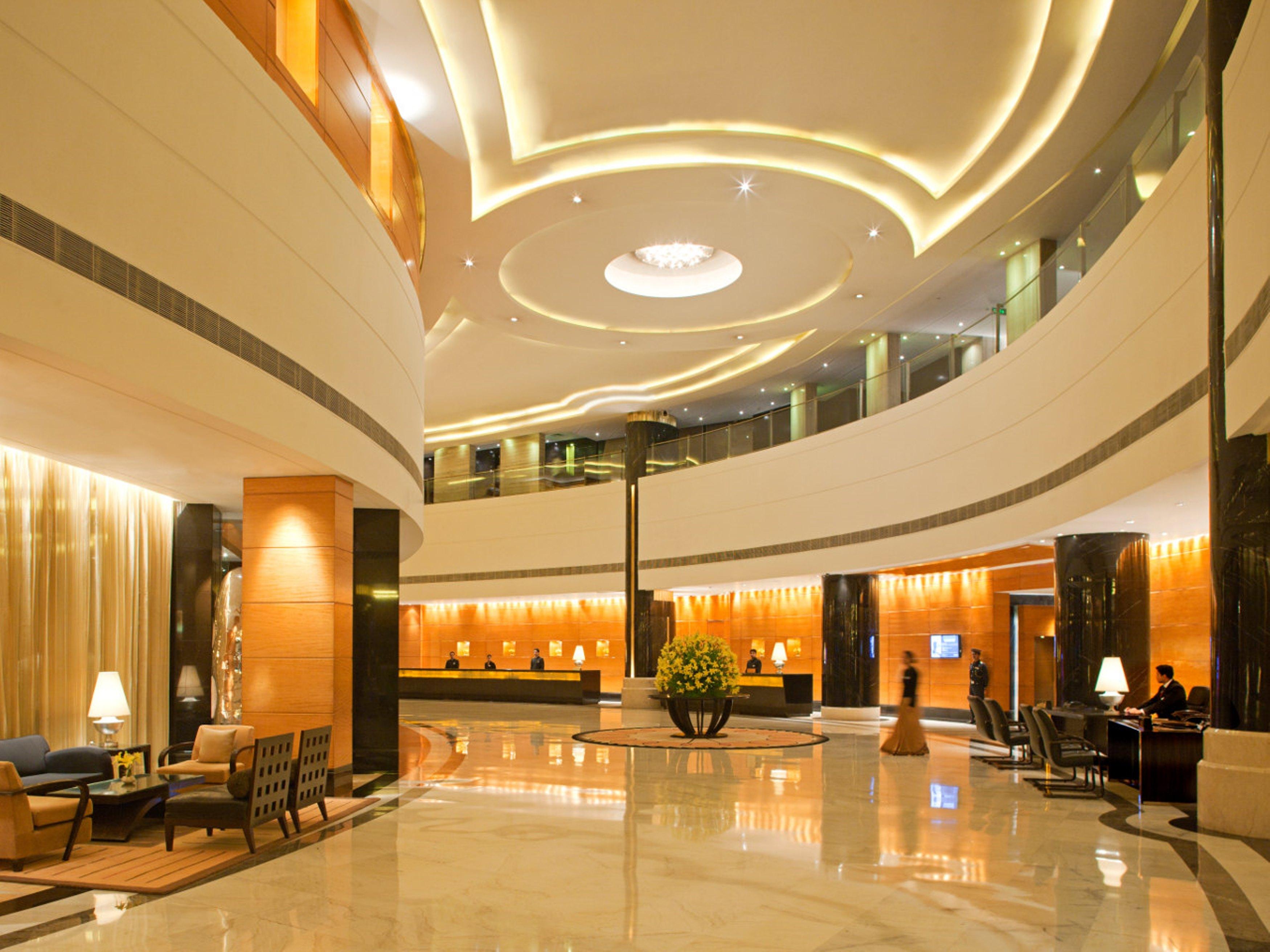 Hotel Radisson Blu Plaza Delhi Airport Nuova Delhi Interno foto The photo shows the interior of a modern hotel lobby. The space features a high ceiling with elegant lighting that enhances the ambiance. There are curved walls and a marble floor that adds to the luxurious feel. The lobby includes reception desks, s