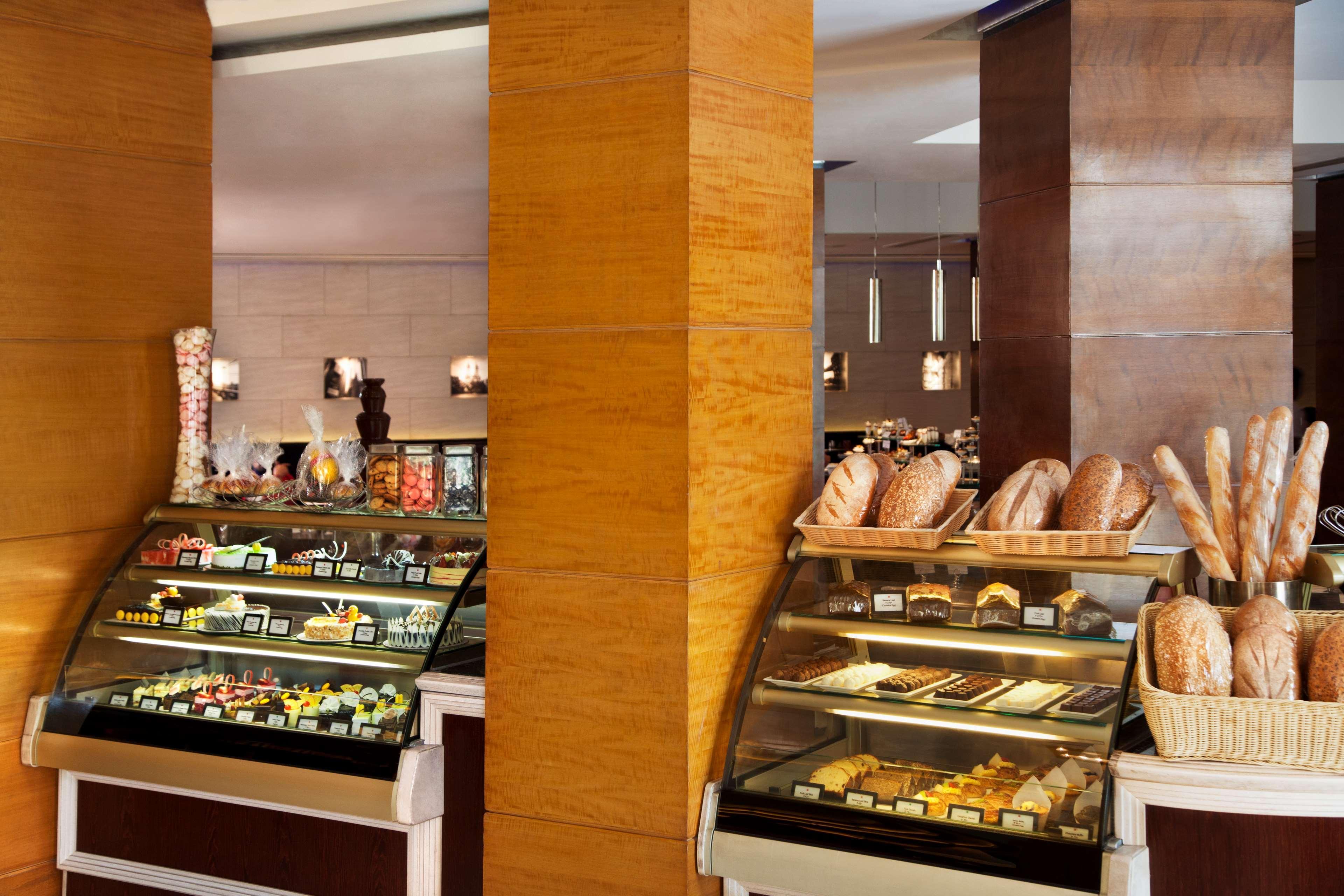 Hotel Radisson Blu Plaza Delhi Airport Nuova Delhi Esterno foto The photo features a bakery display area in a café or restaurant. There are two prominent display cases: one filled with a variety of pastries, cakes, and sweets, and the other showing an assortment of bread, including loaves and baguettes. The inter