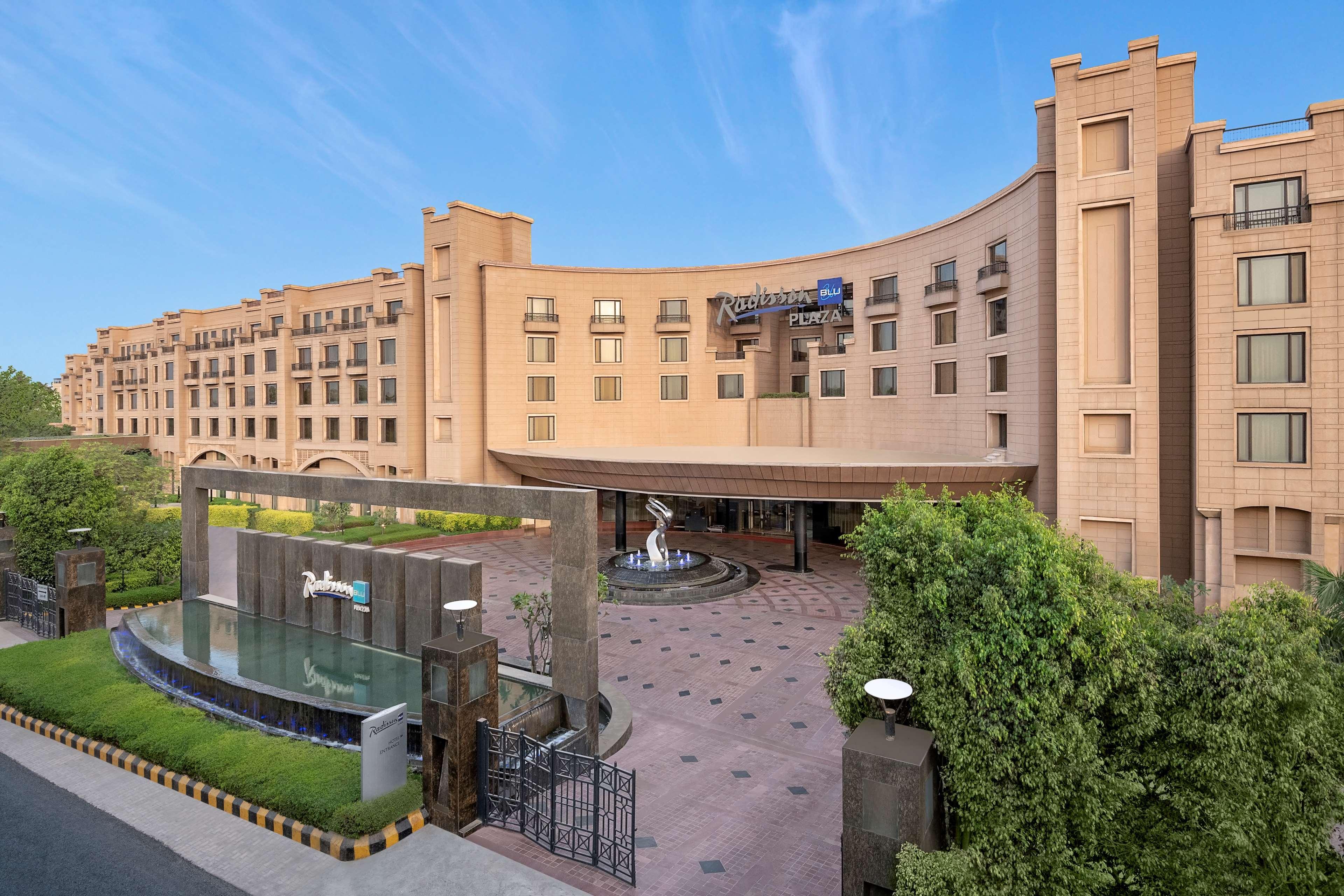 Hotel Radisson Blu Plaza Delhi Airport Nuova Delhi Esterno foto The photo shows the exterior of a modern hotel with a distinctive architectural design. The building features a curved facade and is constructed with a combination of light-colored materials. In the foreground, there is a landscaped area with greener