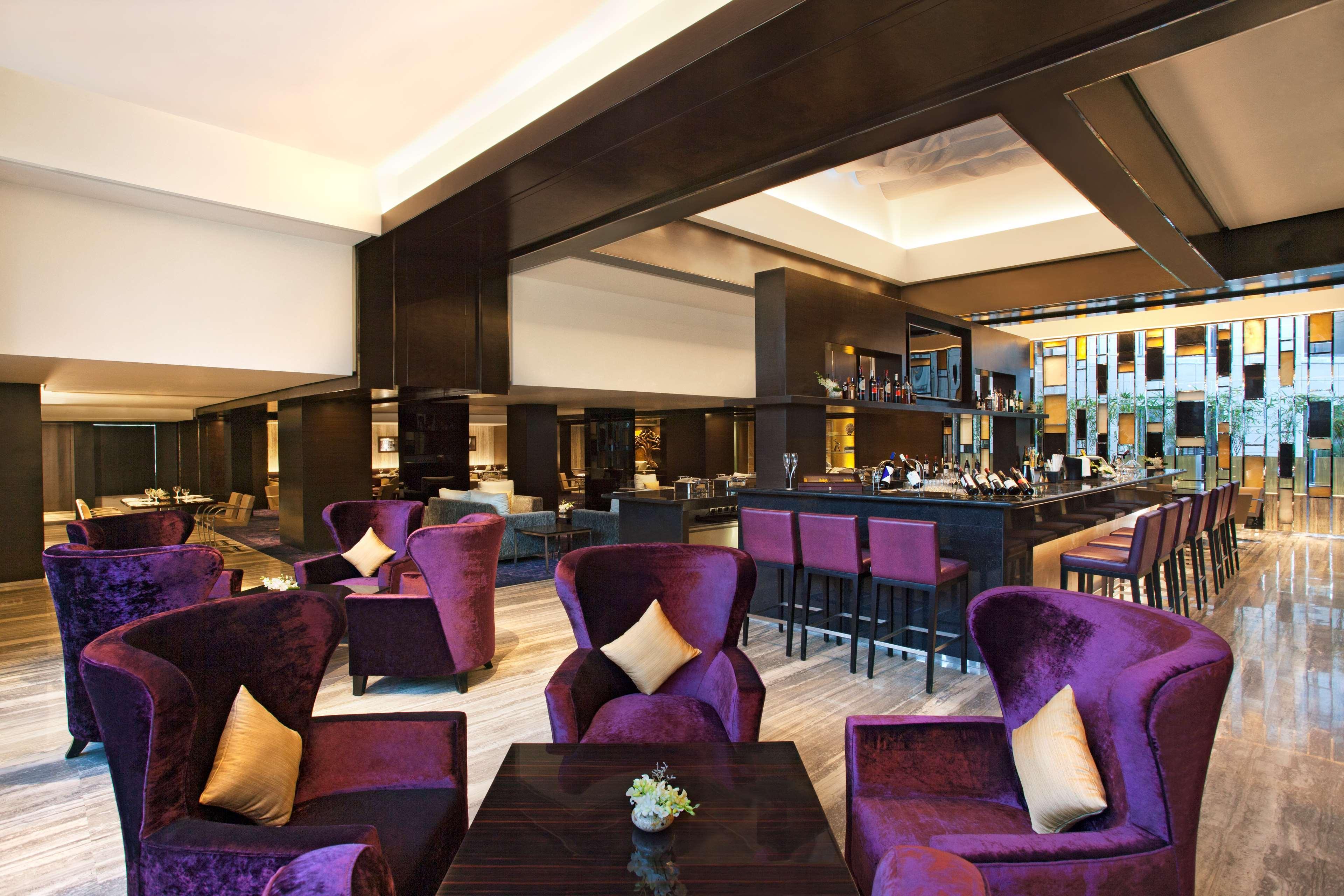 Hotel Radisson Blu Plaza Delhi Airport Nuova Delhi Esterno foto The photo shows a stylish interior of a lounge or bar area. The space features plush purple armchairs with cream-colored cushions arranged around dark wooden tables. In the background, there is a sleek bar with a well-stocked display and seating. The