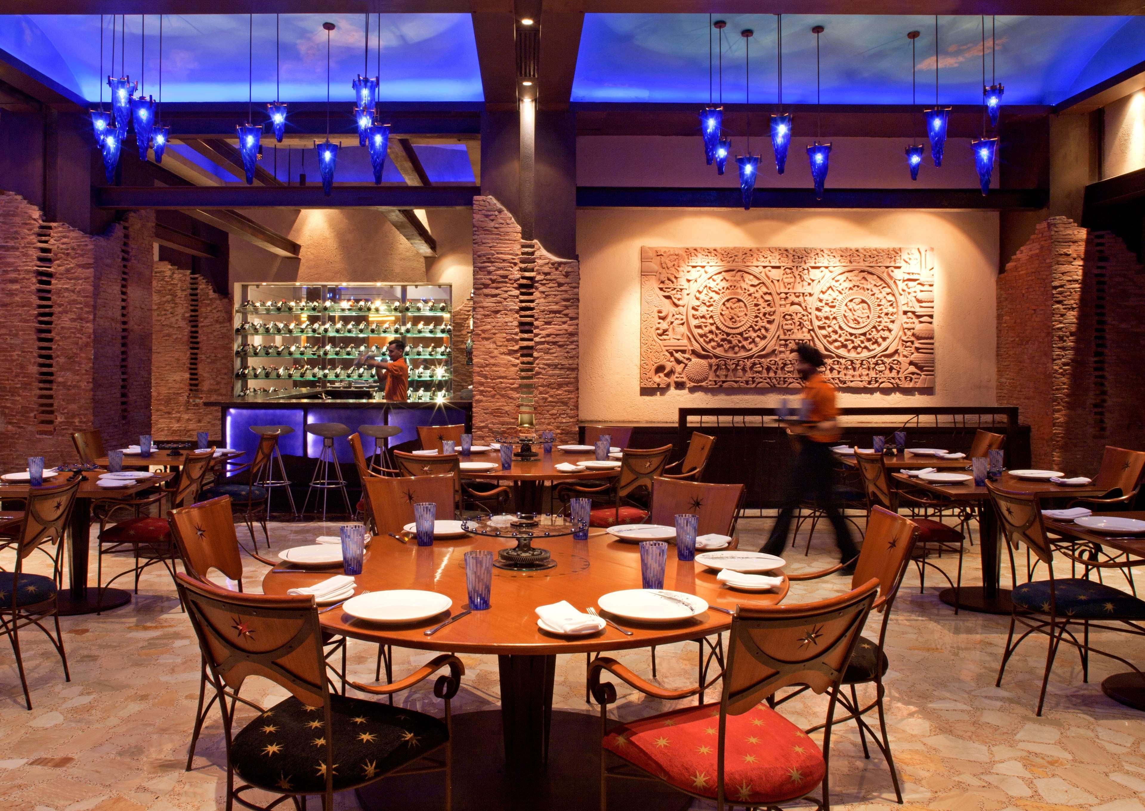 Hotel Radisson Blu Plaza Delhi Airport Nuova Delhi Ristorante foto The photo shows an elegant restaurant interior. There are several round tables arranged with white plates and glassware on them. The chairs have unique designs, featuring a mix of colors and patterns. The walls have a textured appearance, possibly wi