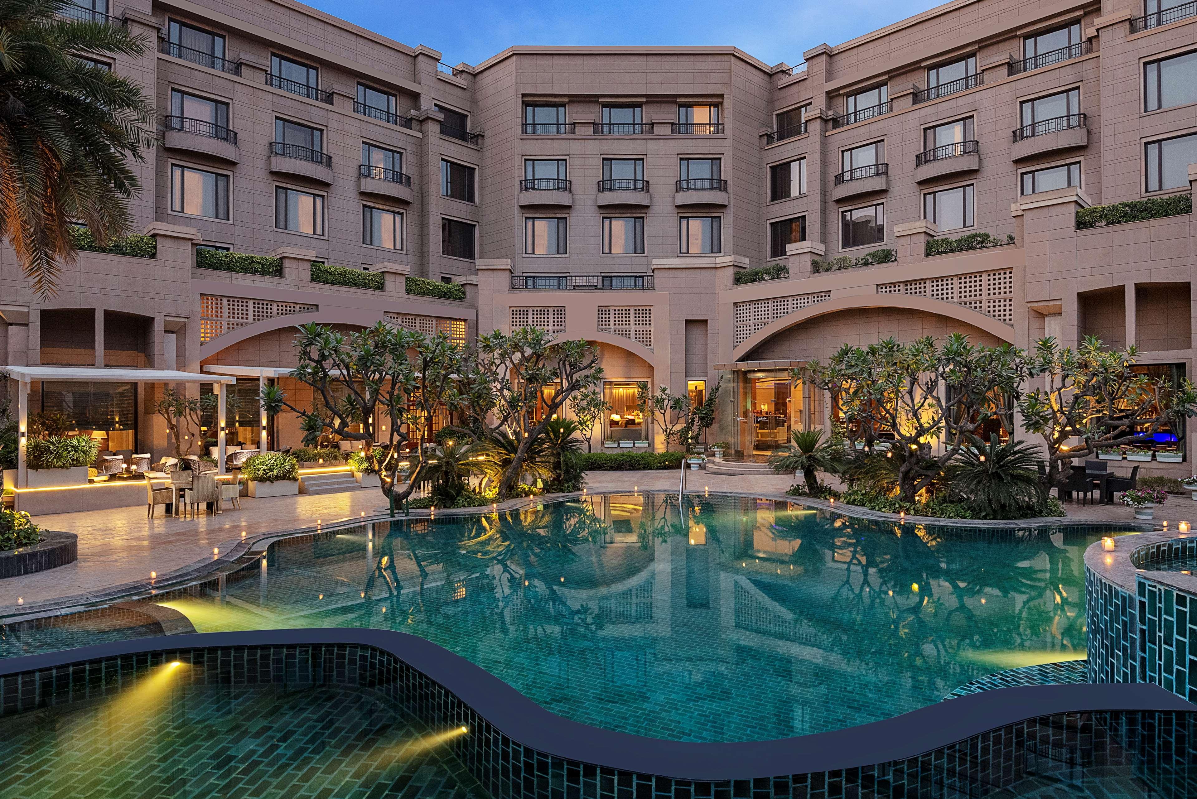 Hotel Radisson Blu Plaza Delhi Airport Nuova Delhi Esterno foto The photo shows a luxurious hotel or resort setting featuring a large swimming pool in the foreground. The pool has a unique, winding shape and is surrounded by lush greenery, including trees and plants. The hotel itself is visible in the background,