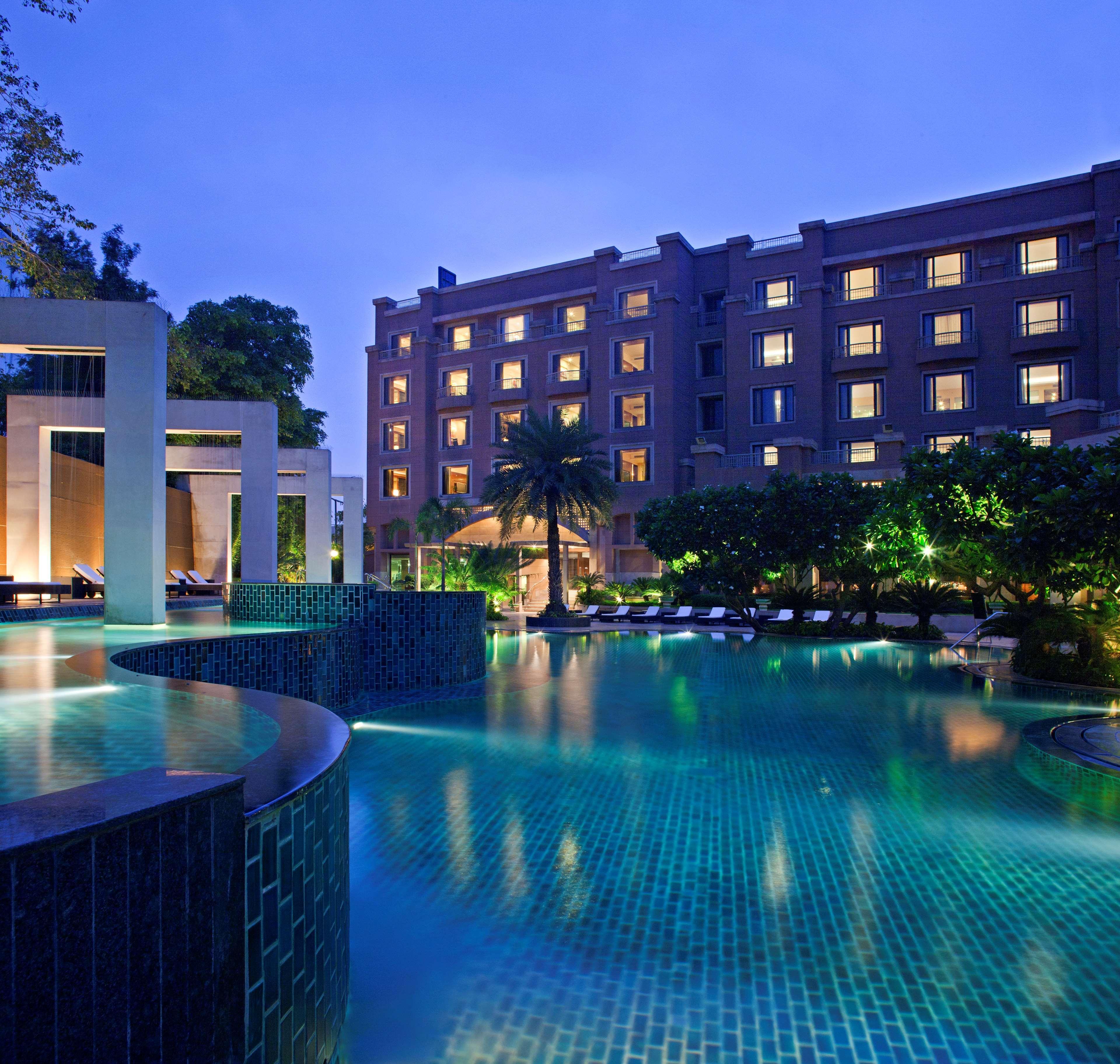 Hotel Radisson Blu Plaza Delhi Airport Nuova Delhi Servizi foto The photo shows a serene outdoor setting featuring a swimming pool illuminated by soft lighting. The pool is surrounded by lush greenery and palm trees, creating a relaxing atmosphere. In the background, there is a stylish hotel or building with mult