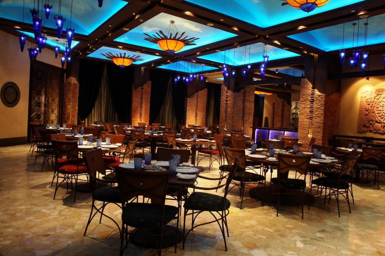 Hotel Radisson Blu Plaza Delhi Airport Nuova Delhi Esterno foto The photo shows a spacious restaurant interior featuring a contemporary design. There are several tables set with plates and utensils, arranged in a neat layout across the room. The ceiling is adorned with an artistic design, illuminated by soft blue
