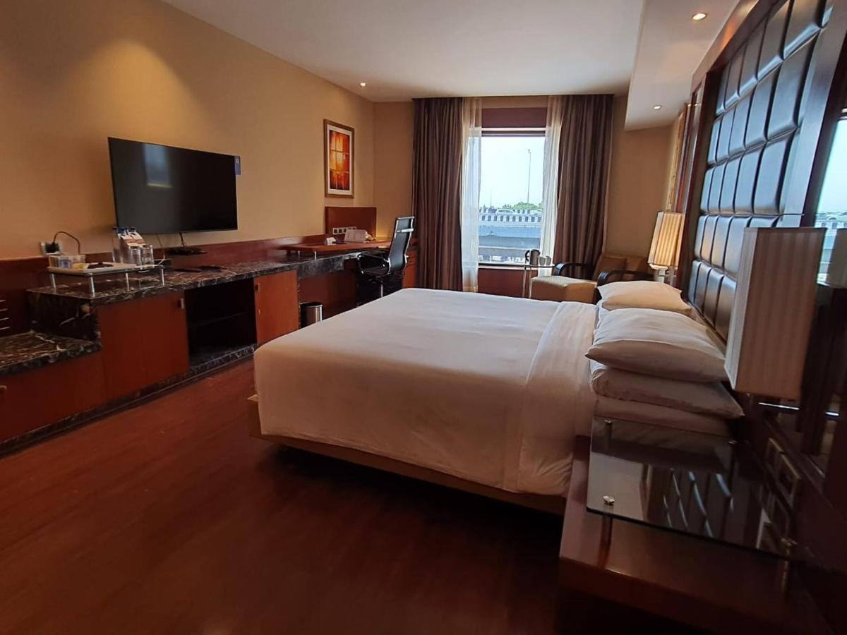 Hotel Radisson Blu Plaza Delhi Airport Nuova Delhi Esterno foto The photo shows a stylish hotel room featuring a large bed with white linens. There is a modern wall-mounted television opposite the bed. To the left, there is a desk area with a chair and additional furniture, including a small bar or kitchenette se