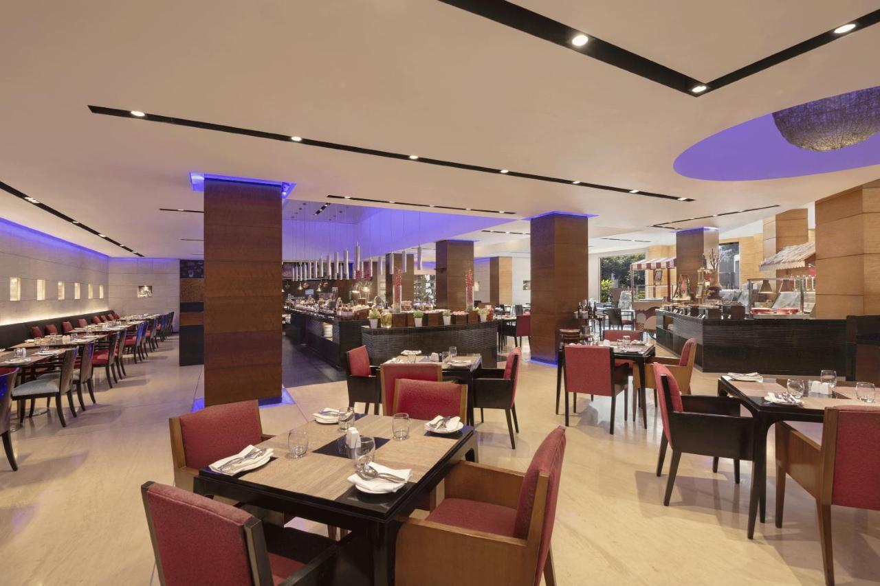 Hotel Radisson Blu Plaza Delhi Airport Nuova Delhi Esterno foto The photo depicts a modern restaurant interior with a spacious layout. There are several tables set for dining, featuring dark wooden surfaces and burgundy chairs. The lighting is bright and includes decorative elements, enhancing the contemporary am