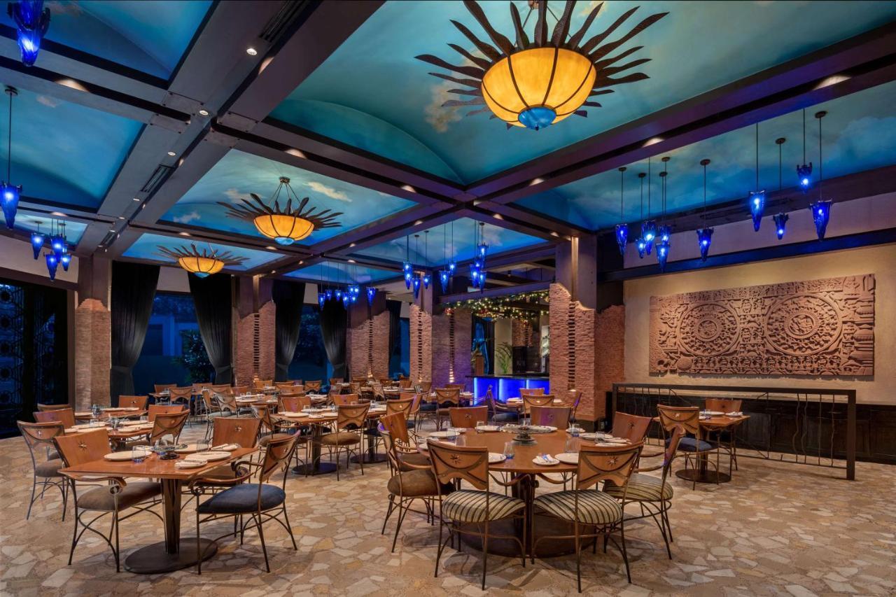 Hotel Radisson Blu Plaza Delhi Airport Nuova Delhi Esterno foto The photo showcases a stylish restaurant interior. It features a spacious dining area with several round tables surrounded by wicker chairs. The ceiling is decorated with a blue hue and adorned with artistic light fixtures resembling suns. There are 