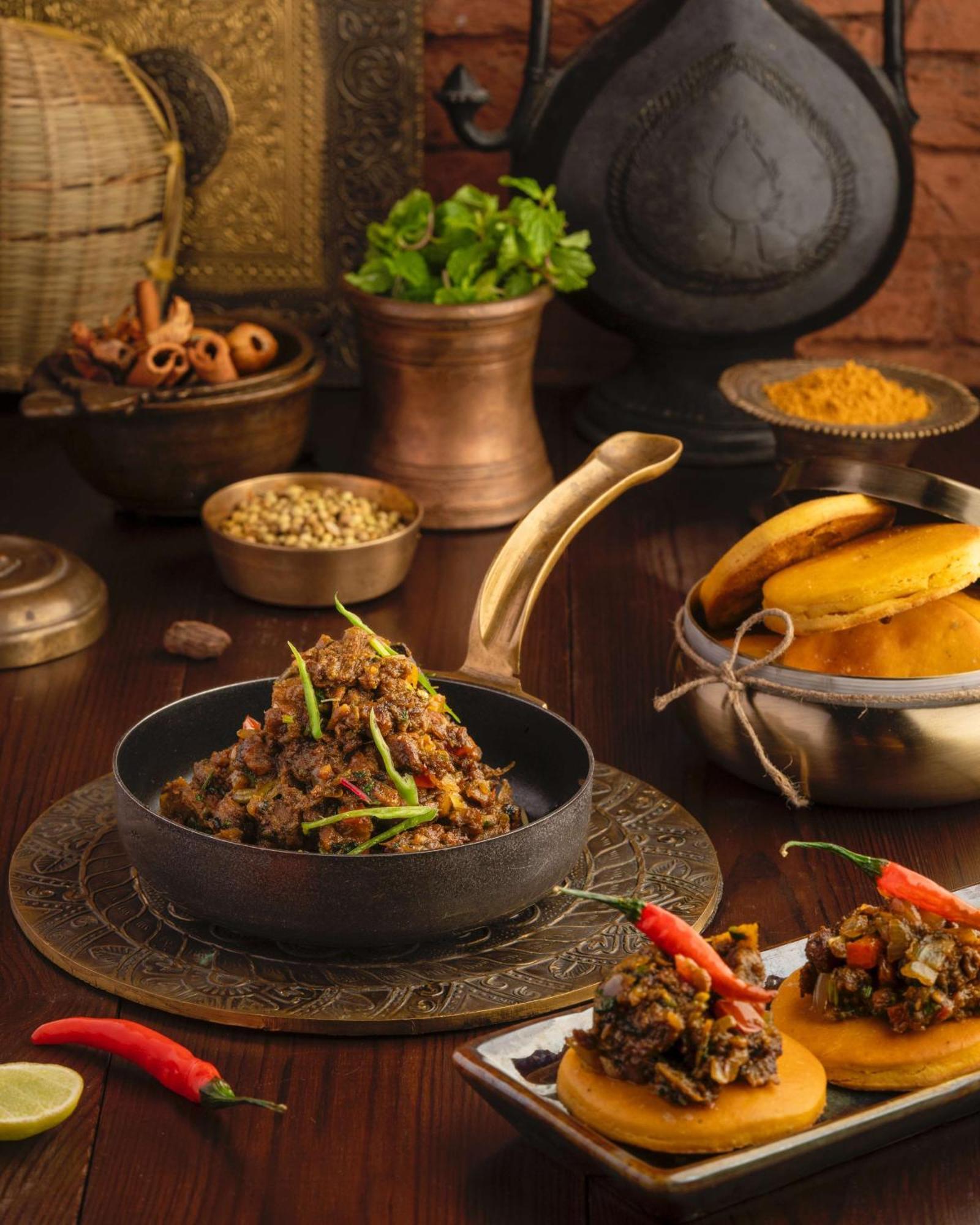 Hotel Radisson Blu Plaza Delhi Airport Nuova Delhi Esterno foto The photo features a traditional meal served in a rustic and decorative setting. In the foreground, there is a dark, shallow pan filled with a flavorful dish, likely meat or vegetables, garnished with green chilies. Next to it, a silver bowl contains