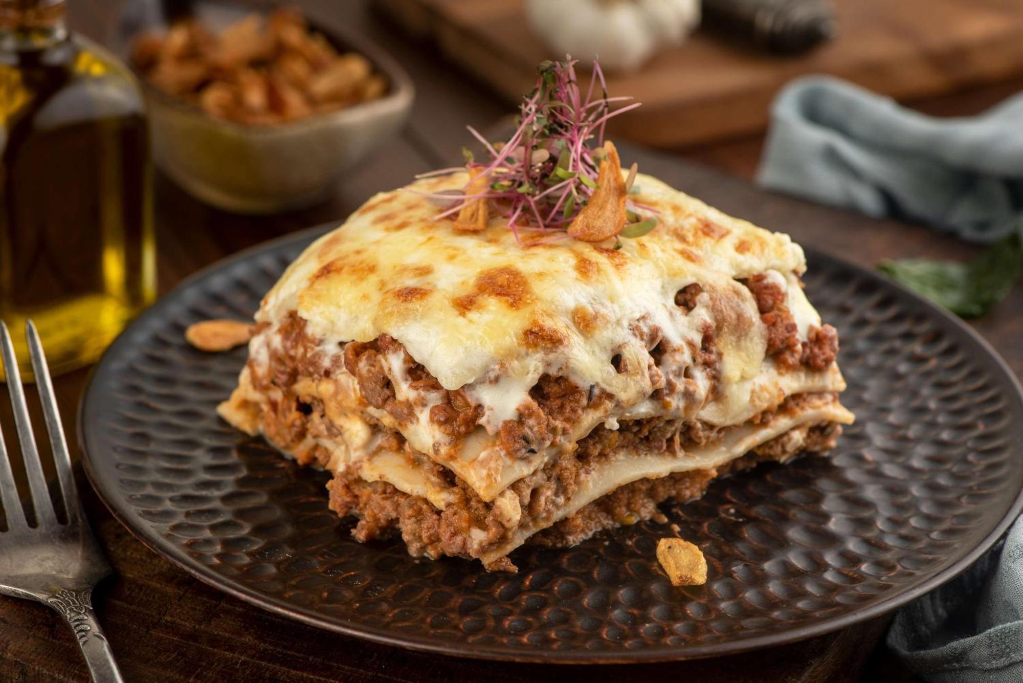 Hotel Radisson Blu Plaza Delhi Airport Nuova Delhi Esterno foto The photo shows a delicious serving of lasagna on a dark, textured plate. The lasagna is layered with pasta, meat, and cheese, topped with a golden, melted cheese layer. It is garnished with small sprouts or herbs for decoration. In the background, t