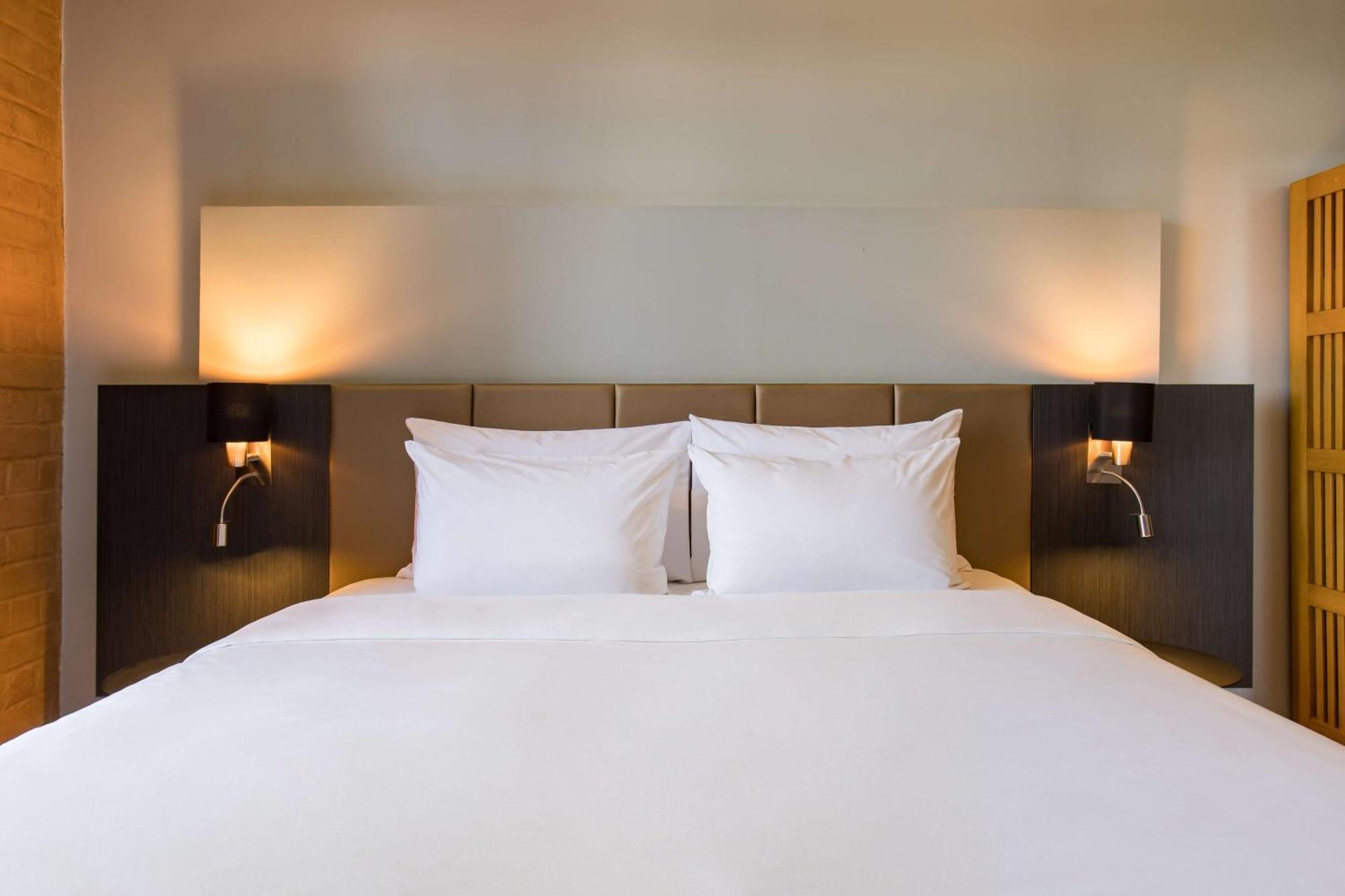 Hotel Radisson Blu Plaza Delhi Airport Nuova Delhi Esterno foto The photo shows a modern, neatly made bed with white bedding and two fluffy, white pillows. The bed is positioned against a light-colored wall, and there are two bedside tables on either side, each featuring a stylish black lamp. The overall aestheti