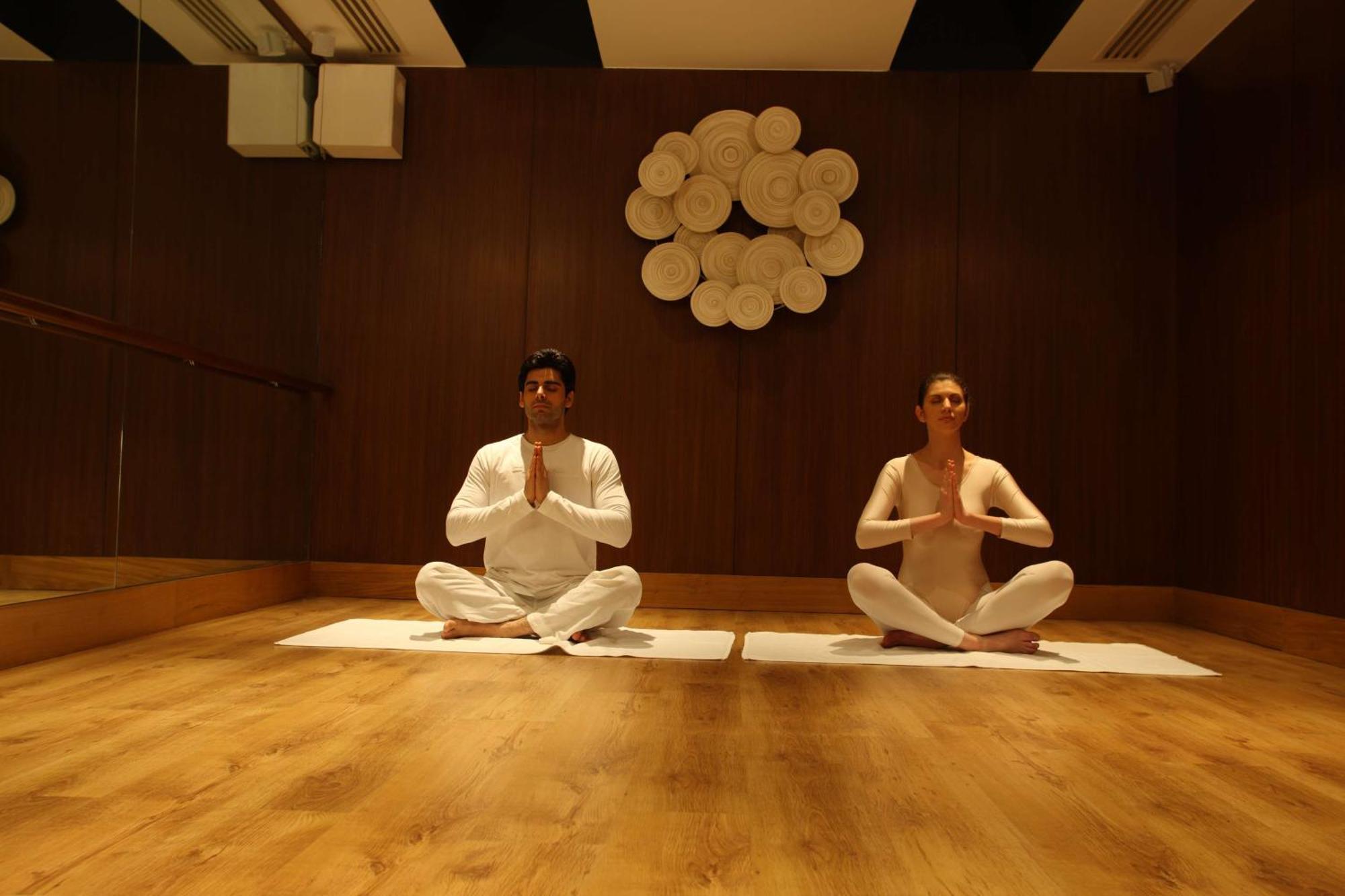 Hotel Radisson Blu Plaza Delhi Airport Nuova Delhi Esterno foto The photo shows two individuals seated in a meditation or yoga pose on mats in a serene indoor environment. They are facing each other and have their hands brought together in a prayer position at their chests. The room features wooden flooring and a
