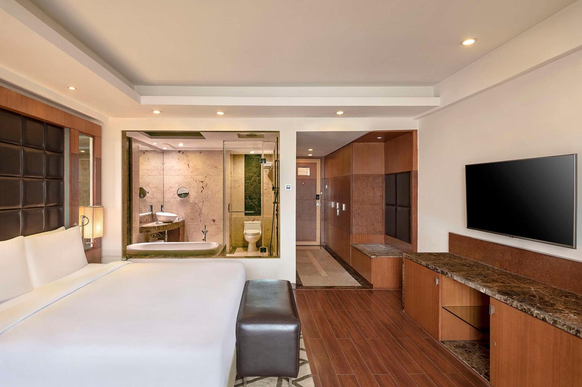 Hotel Radisson Blu Plaza Delhi Airport Nuova Delhi Esterno foto The photo shows a modern hotel room featuring a bed with white linens and a dark-colored ottoman at the foot. To one side, there is a large flat-screen TV mounted on the wall, along with wooden cabinetry and a granite surface for additional storage o