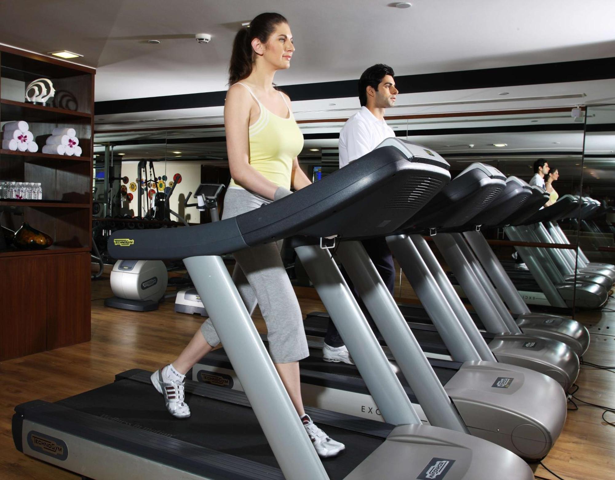 Hotel Radisson Blu Plaza Delhi Airport Nuova Delhi Esterno foto The photo shows a gym scene with two individuals using treadmills. The woman is in the foreground, walking on a treadmill while wearing a yellow tank top and light gray pants. The man is in the background, also on a treadmill, dressed in a white shir