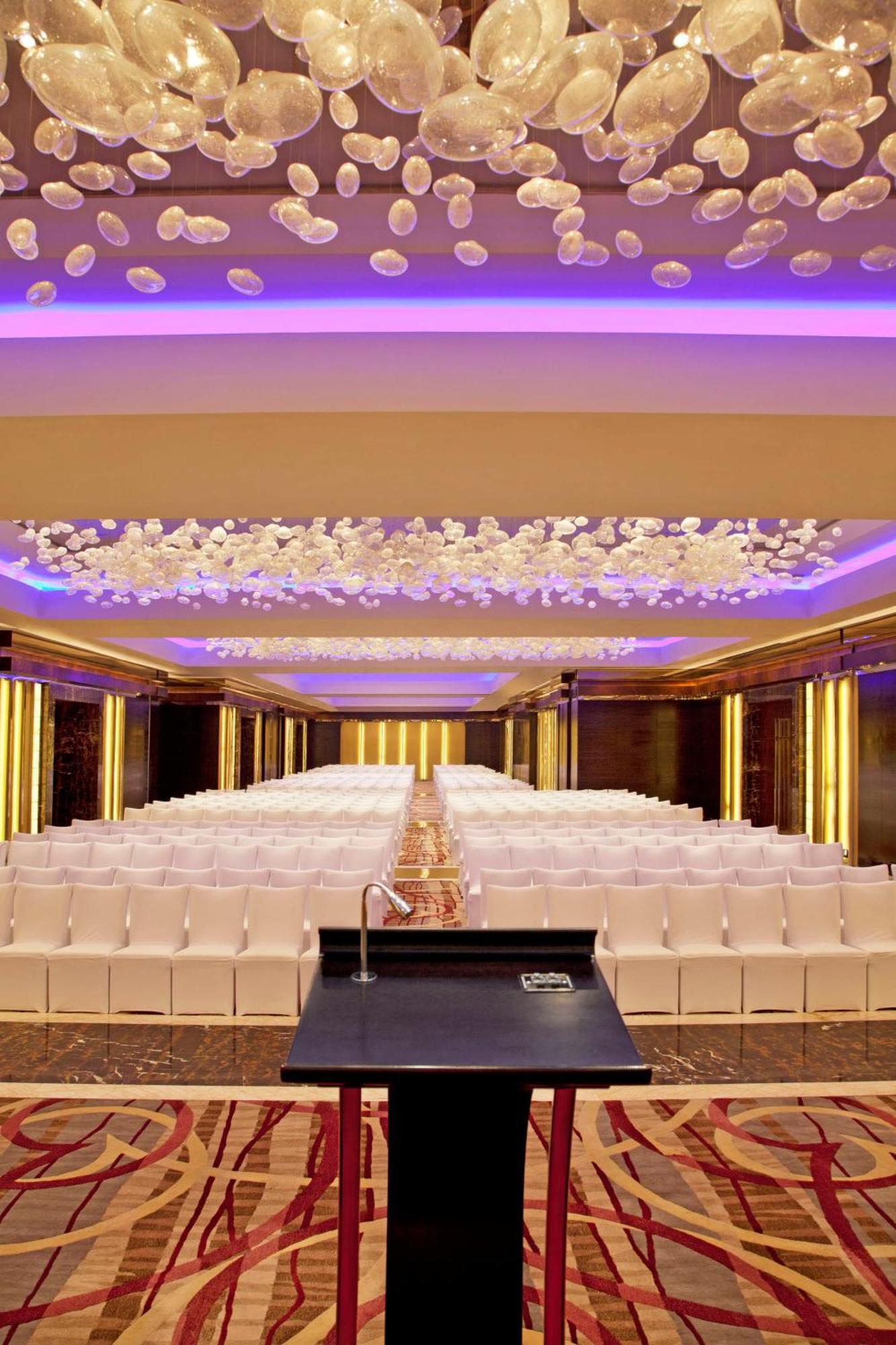 Hotel Radisson Blu Plaza Delhi Airport Nuova Delhi Esterno foto The image shows an elegantly designed indoor event space. It features rows of white chairs arranged neatly for an audience. The ceiling is adorned with decorative elements, possibly flowers, illuminated by soft purple lighting that adds a sophisticat