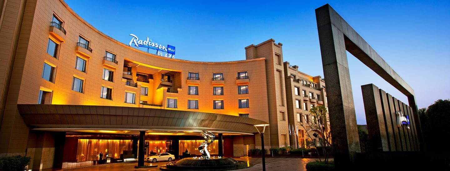 Hotel Radisson Blu Plaza Delhi Airport Nuova Delhi Esterno foto The photo shows the exterior of the Radisson Blu hotel, featuring a modern architectural design. The building is well-lit, with warm lighting highlighting its structure during what appears to be dusk. There is a large entrance area with a canopy, and